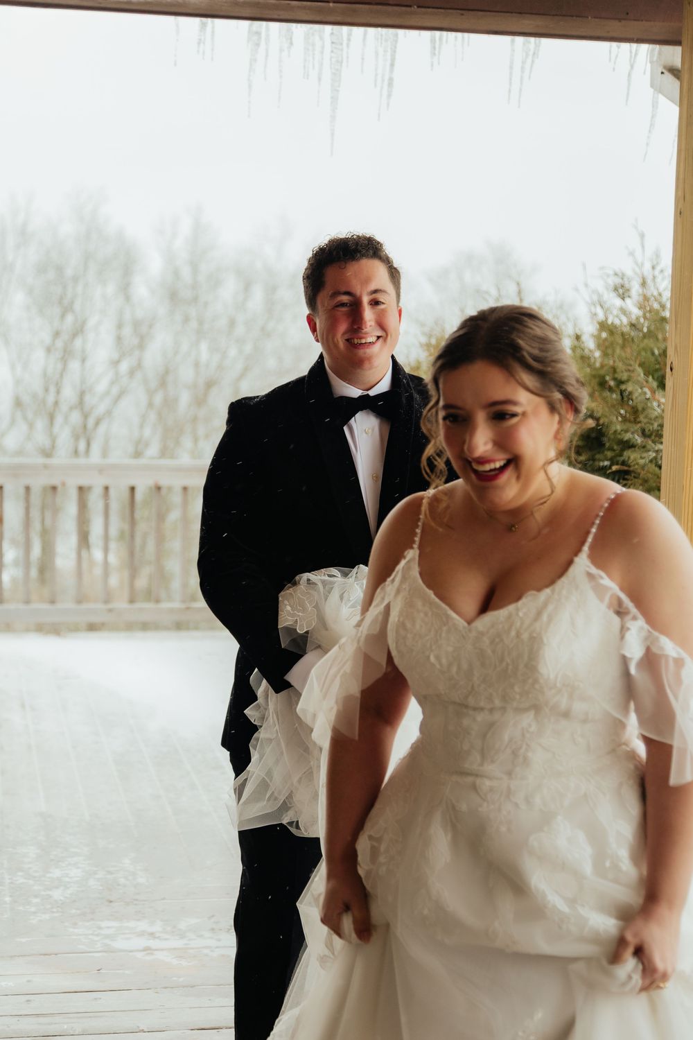 Laughing bride in lace wedding gown and groom in tuxedo stepping inside from a snowy winter wedding day.