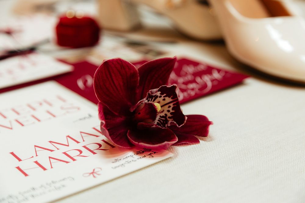 Deep red orchid resting on wedding invitation suite with nude heels and velvet ring box.
