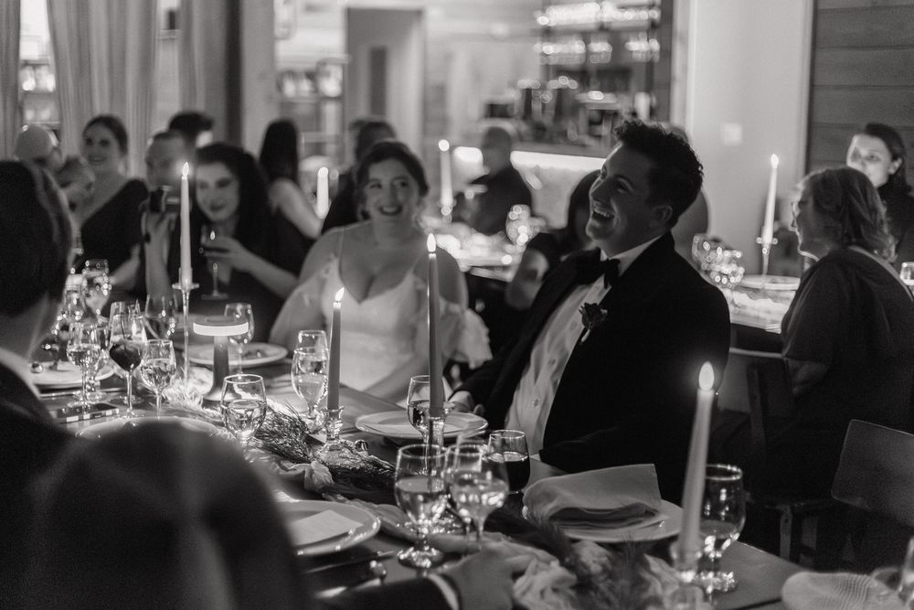 Black and white photo of a bride and groom laughing at their wedding reception dinner table.
