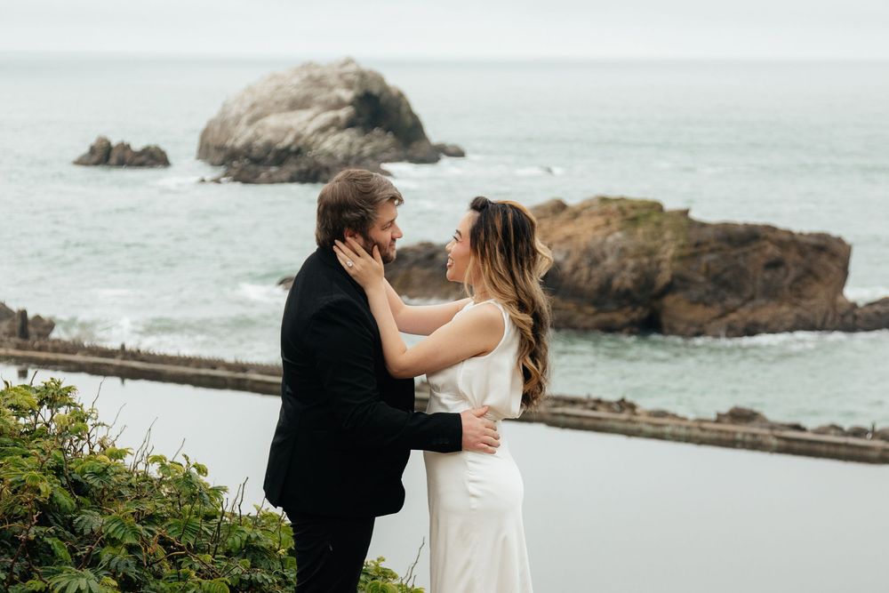 Couple in wedding attire sharing a tender embrace with dramatic rocky Pacific Ocean coastline in the background.