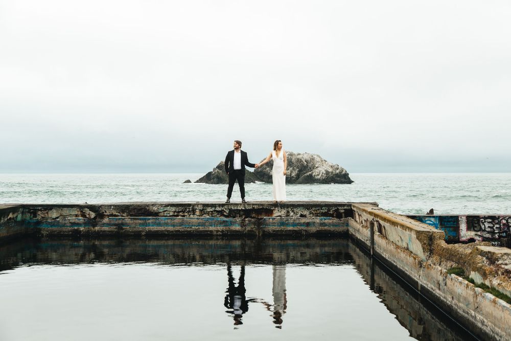 Bride and groom holding hands on a weathered coastal pier with ocean rocks and cloudy sky reflected below.