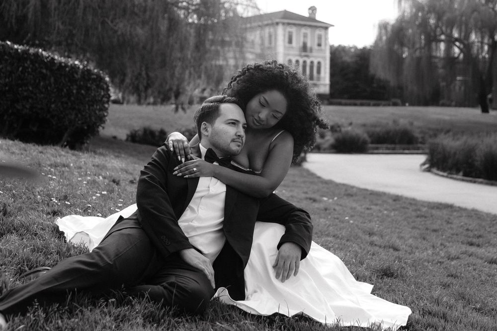 Black and white photo of newlyweds relaxing on grass, bride draped over groom with elegant estate in background.