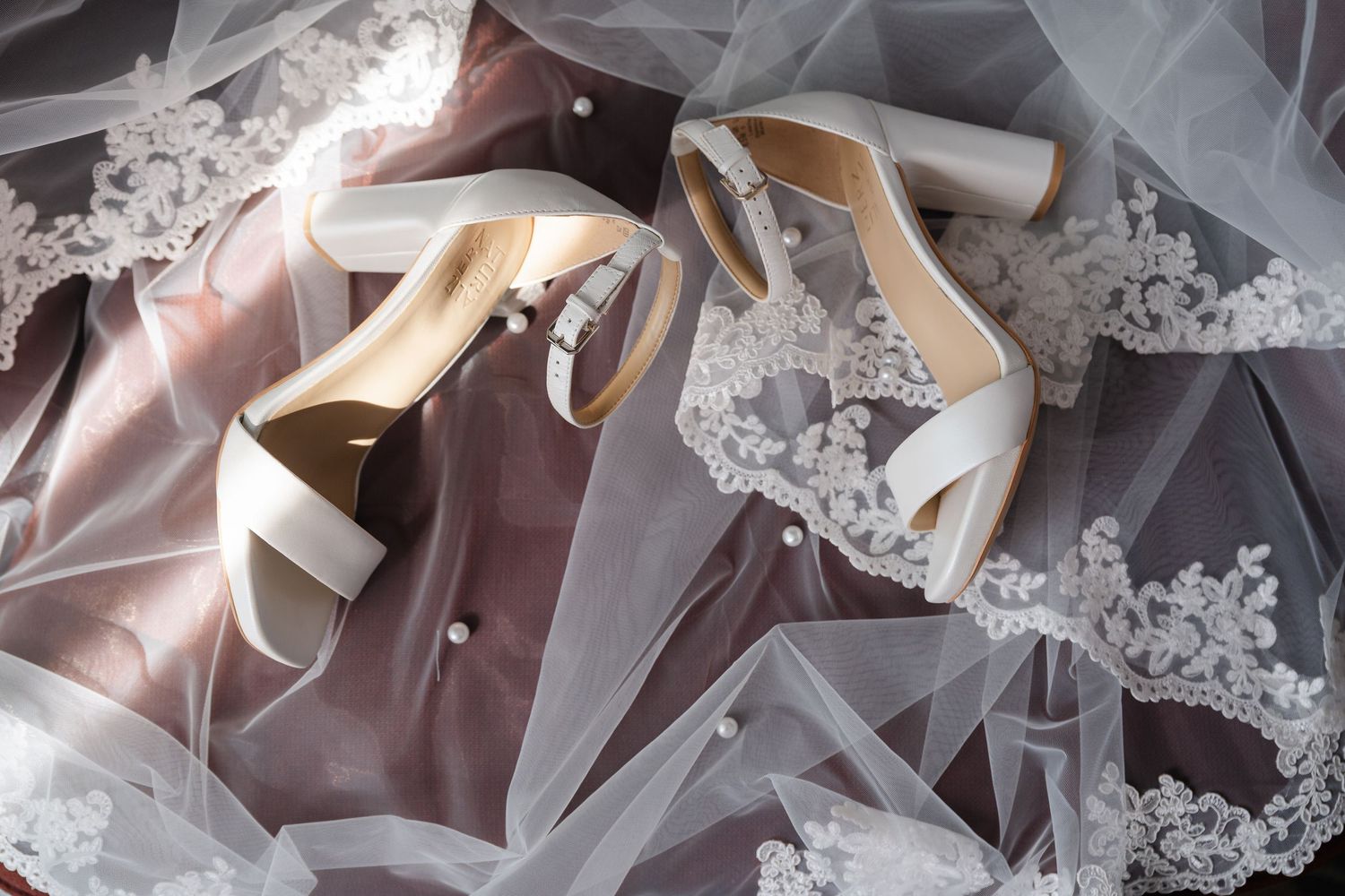 Elegant white wedding heels displayed on delicate lace veil and blush fabric.