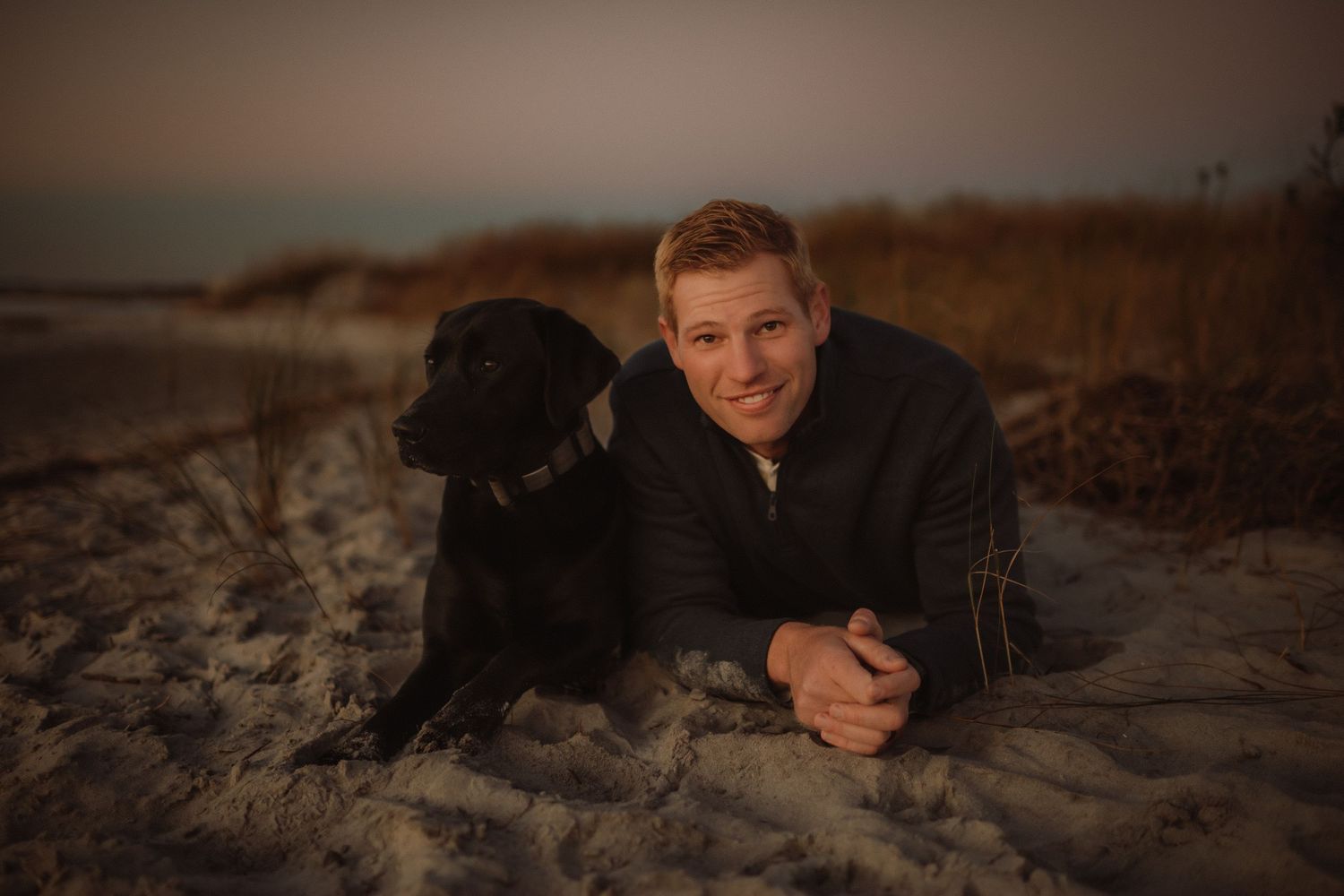 Someone in a black sweater lies on a sandy beach at sunset with their black Labrador retriever companion.