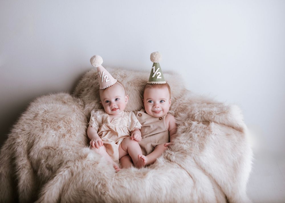 Twin babies wearing party hats sit together on fluffy cream-colored blanket.
