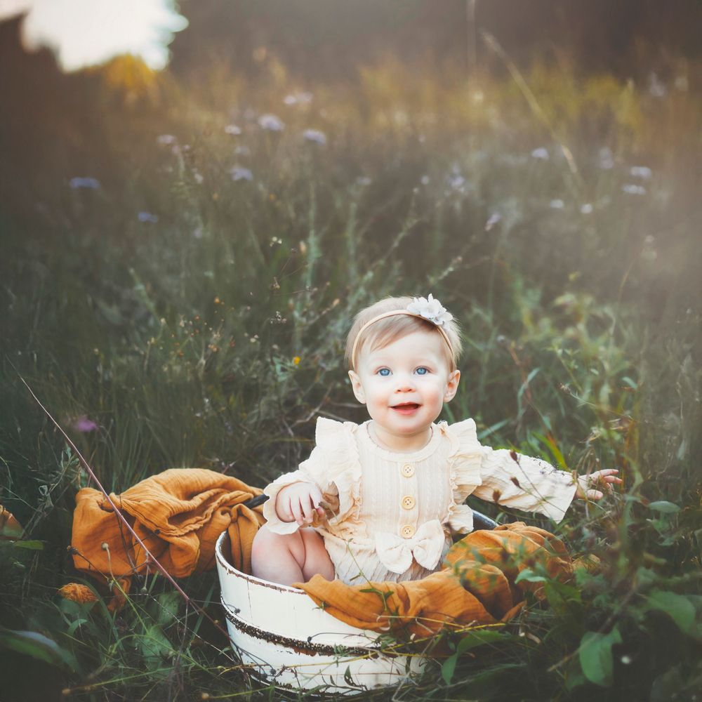 Baby in white dress sits in rustic basket surrounded by wildflowers at sunset.