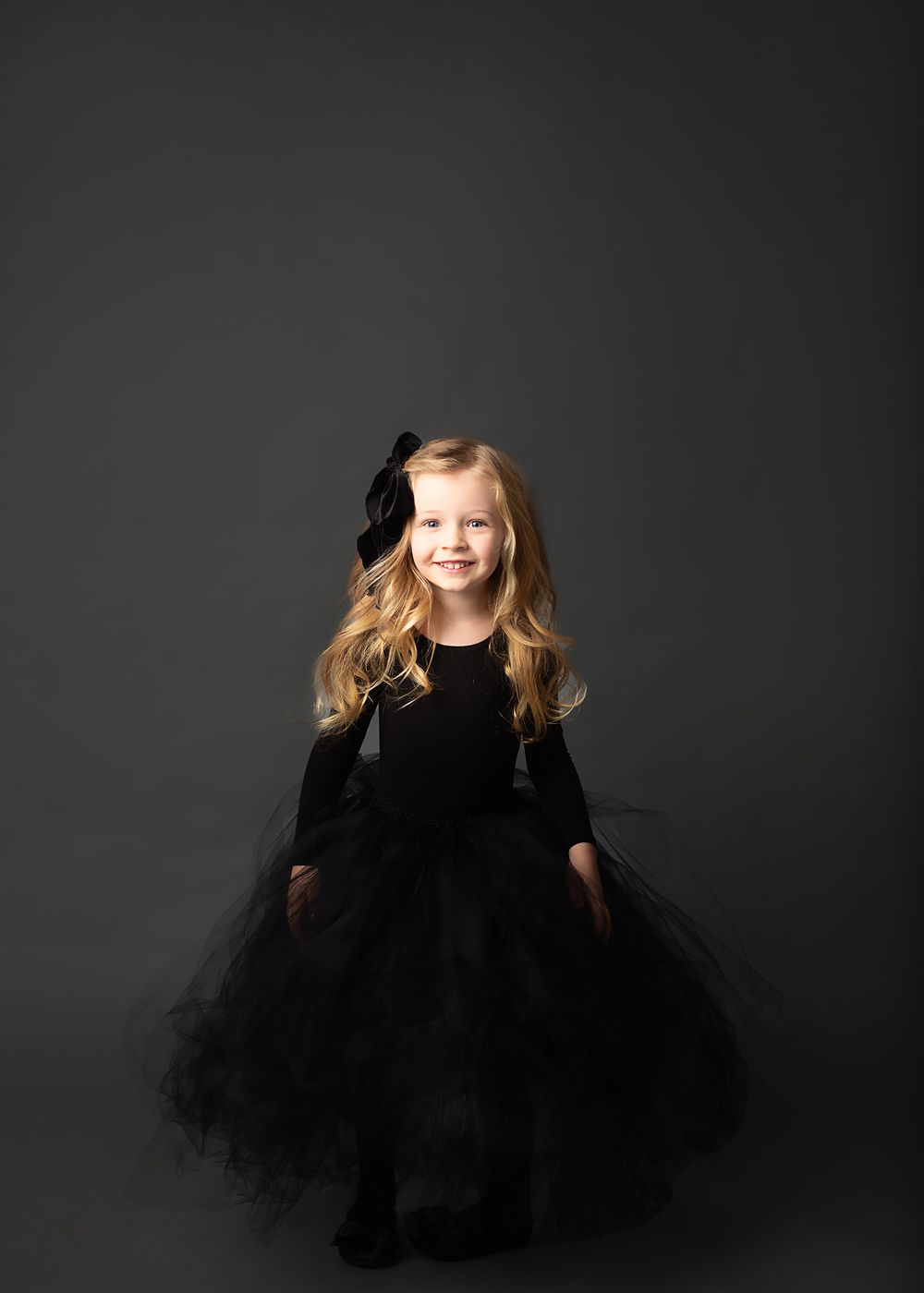 Child wearing black tulle dress poses against dark background in studio portrait.