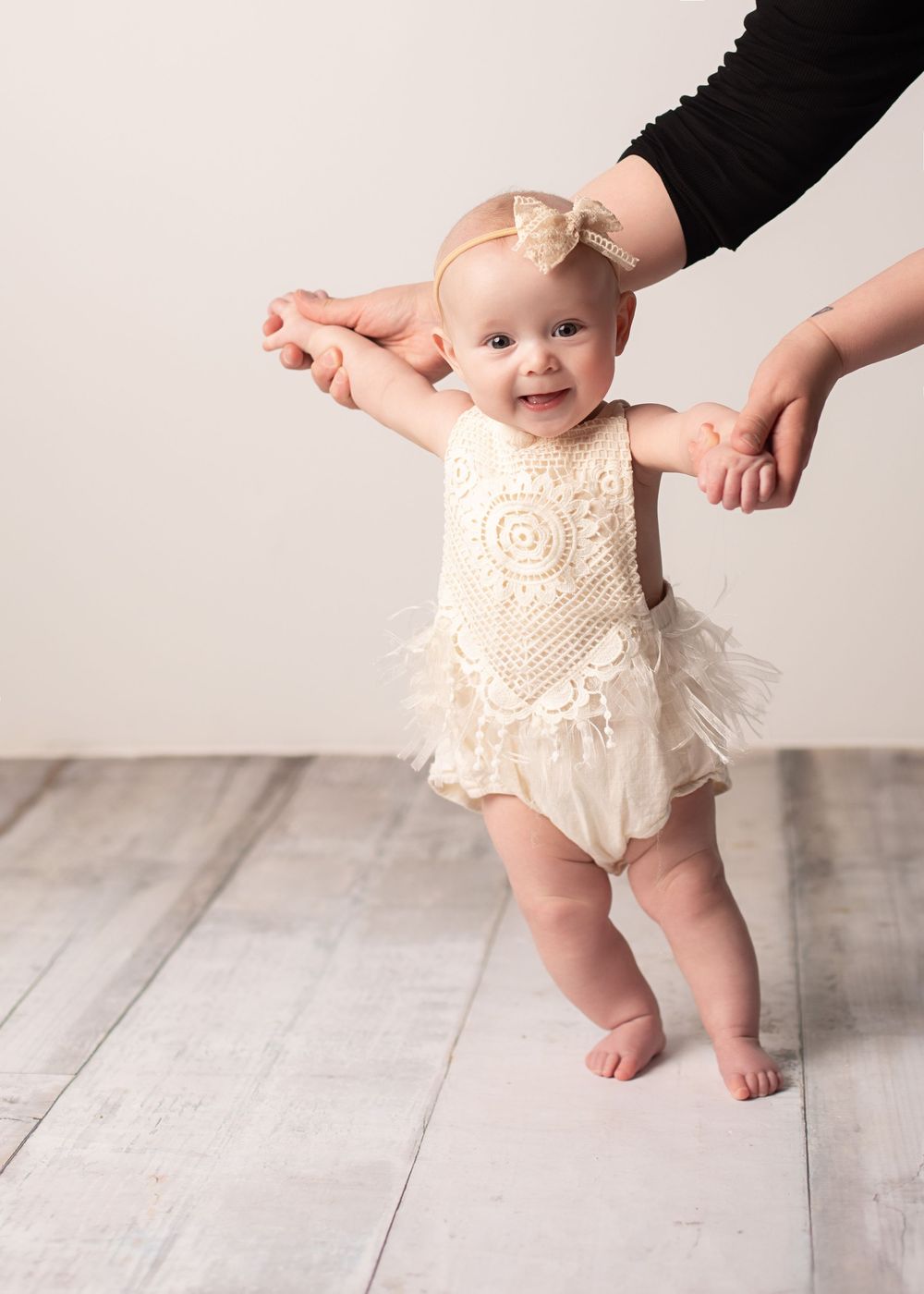A baby in a white lace dress takes first steps while supported by adult hands on a light wooden floor.