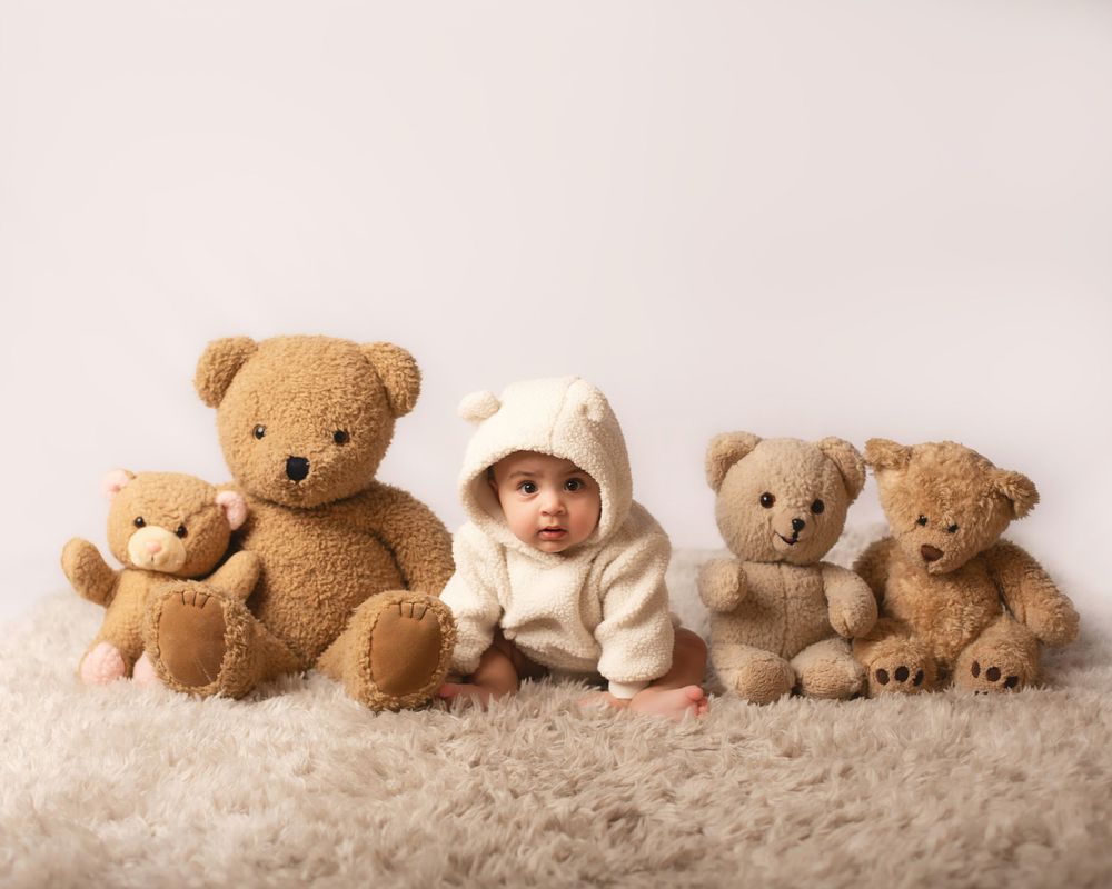 A collection of soft brown teddy bears arranged on a plush carpet with a baby wearing a cozy white bunny outfit in the middle.
