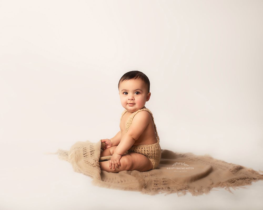 Baby sitting on tan textured blanket in minimalist studio setting.