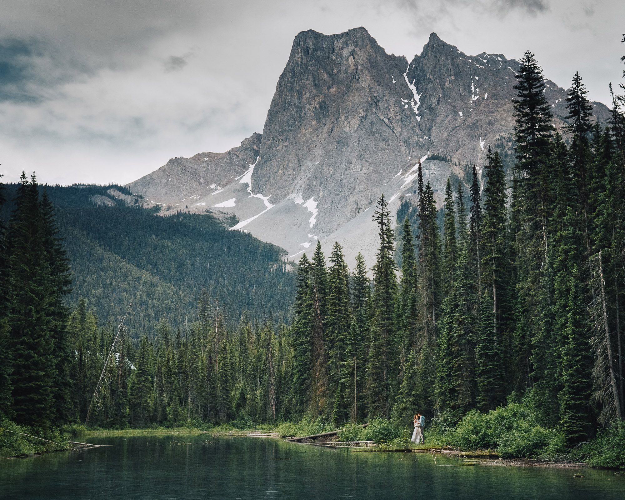 Dramatic mountain landscape with evergreen forest and snow-capped peaks reflecting in a serene lake.
