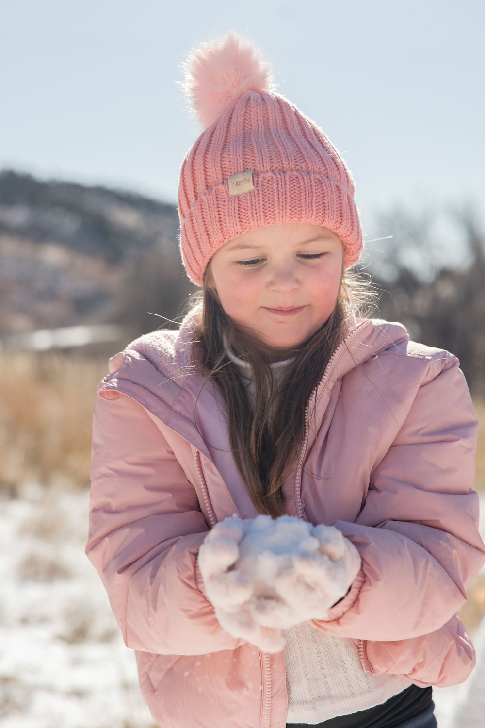 Person wearing pink winter hat and coat playing with snow on a bright winter day.