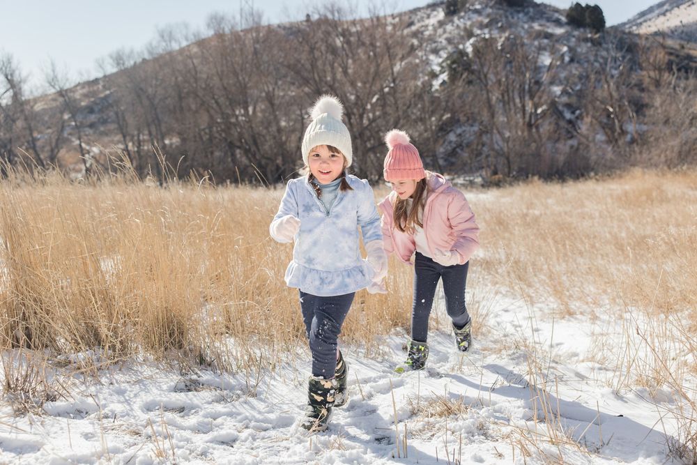 Two children run through snow-covered grass with mountains visible behind them.