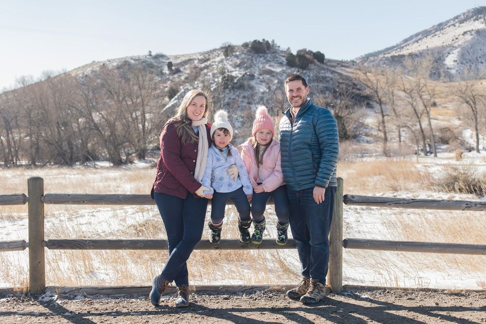 A group poses together on a winter day by a wooden fence with snow-covered mountains in the background.