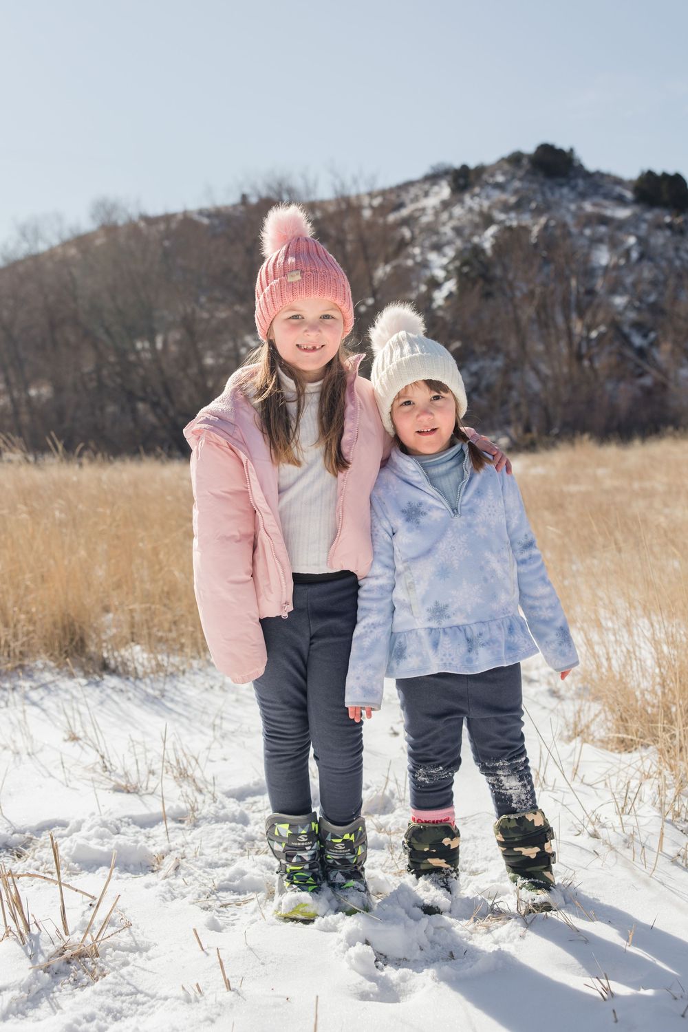 Two children in winter coats and knit hats stand together in a snowy field with mountains in the background.