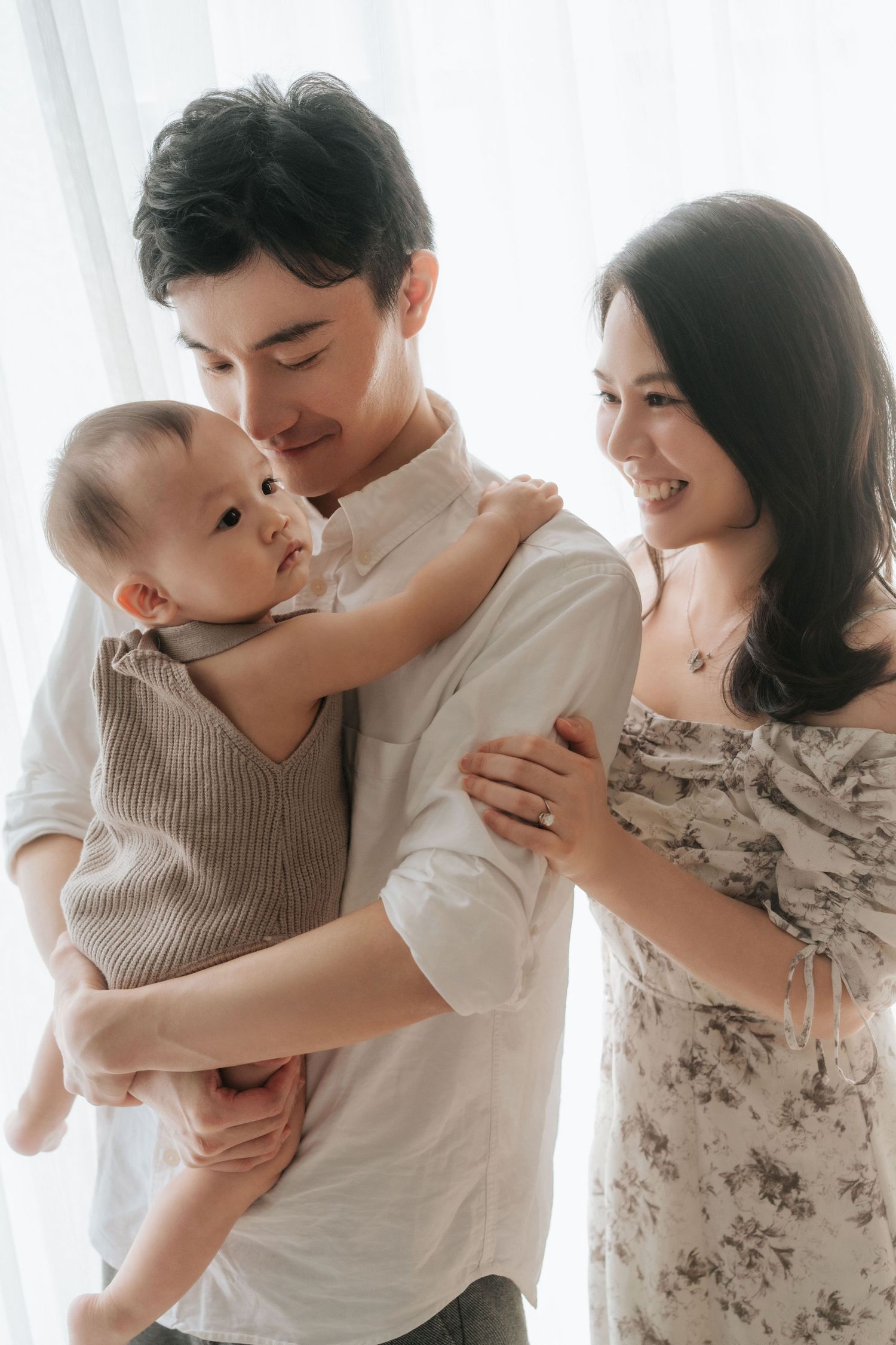 Happy Asian family with father holding baby while smiling mother stands beside them near bright window.幸福的亞洲家庭，父親抱著嬰兒，微笑的母親站在明亮的窗邊。
