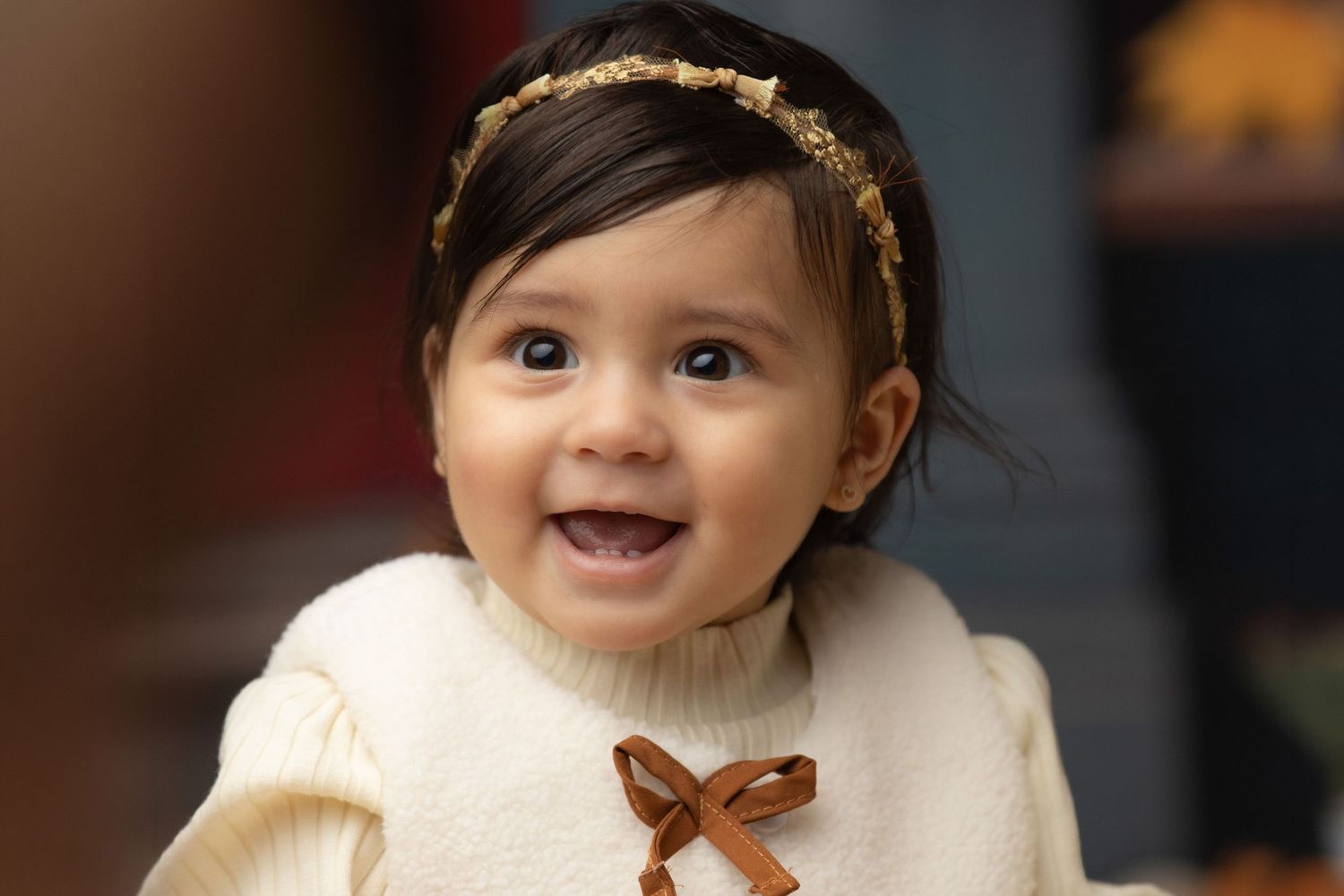 Baby wearing a white knit sweater and gold headband smiles joyfully at the camera.
