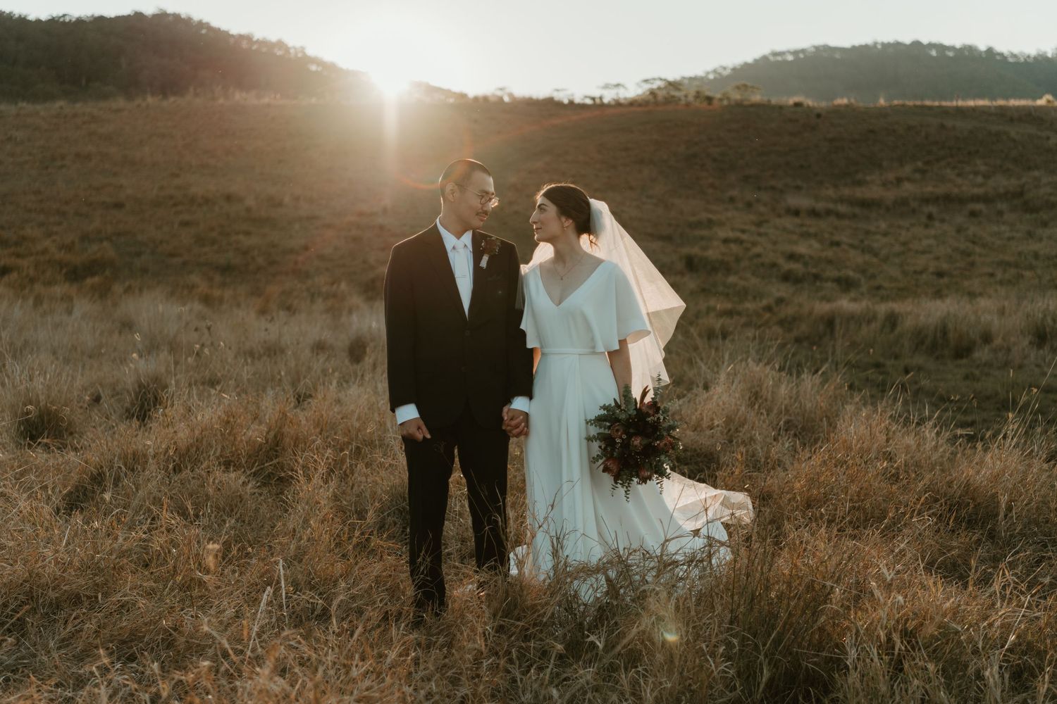 Bride and groom stand in golden sunlit field, bride in white gown with veil, groom in black suit, sharing a romantic moment.