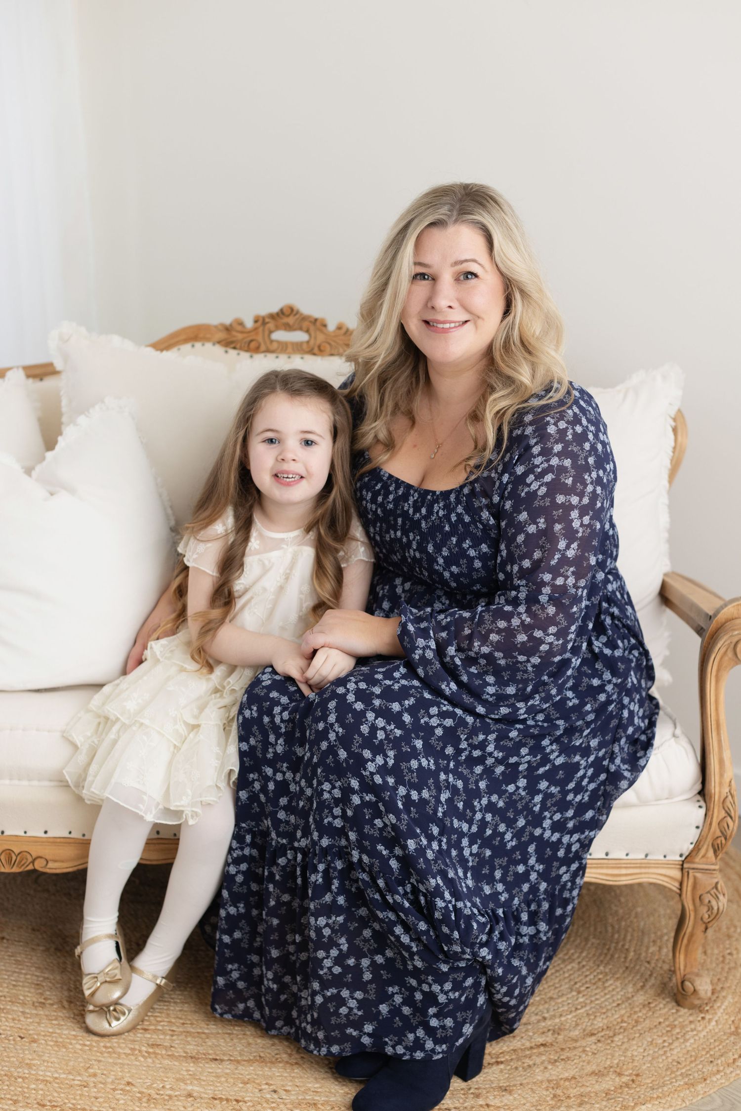 A parent and child sit together on an elegant cream-colored vintage sofa wearing coordinating blue and white dresses.