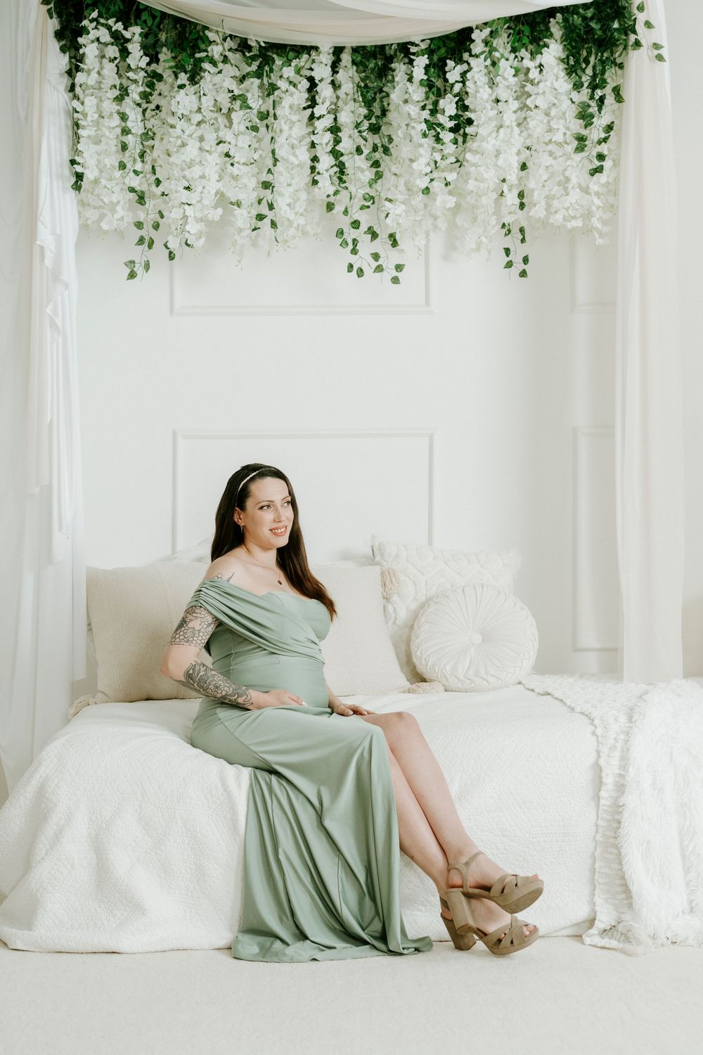 Maternity photoshoot in white studio with hanging greenery and sage green dress on white couch.