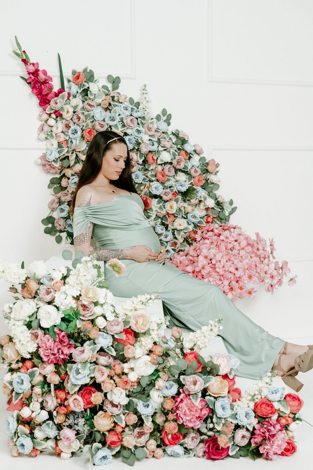 A maternity photoshoot features a flowing mint green dress surrounded by a lush arrangement of pink and coral roses.
