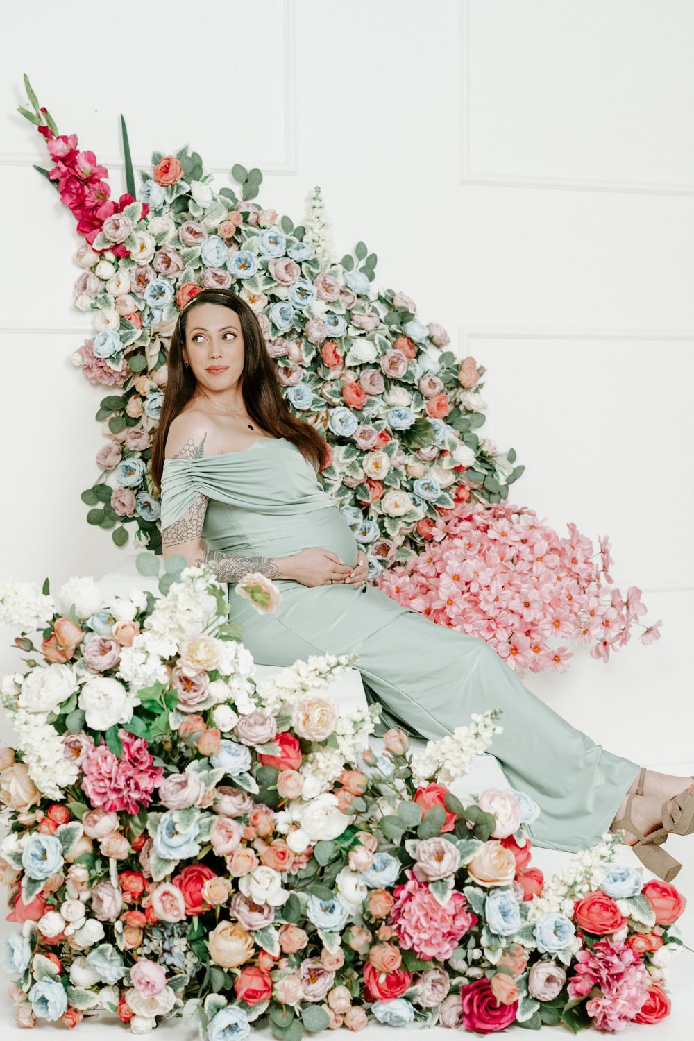 Maternity photo shoot featuring a mint green dress against a romantic backdrop of pink and white floral arrangements.