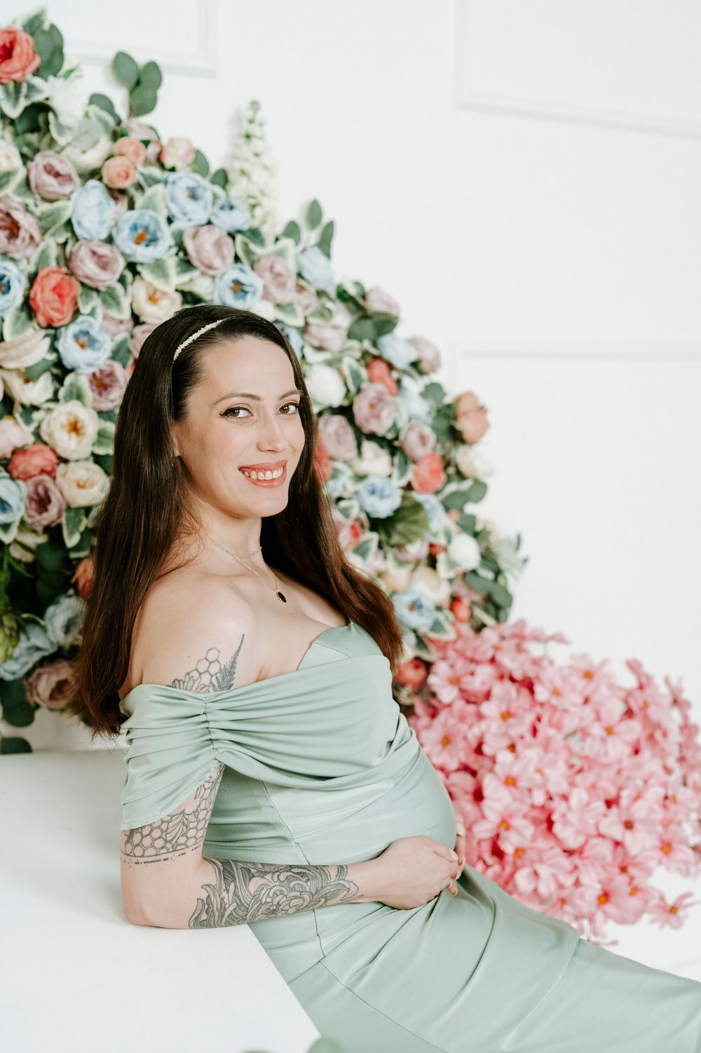 A woman in a mint green dress poses against a backdrop of pink and coral floral arrangements in a studio setting.