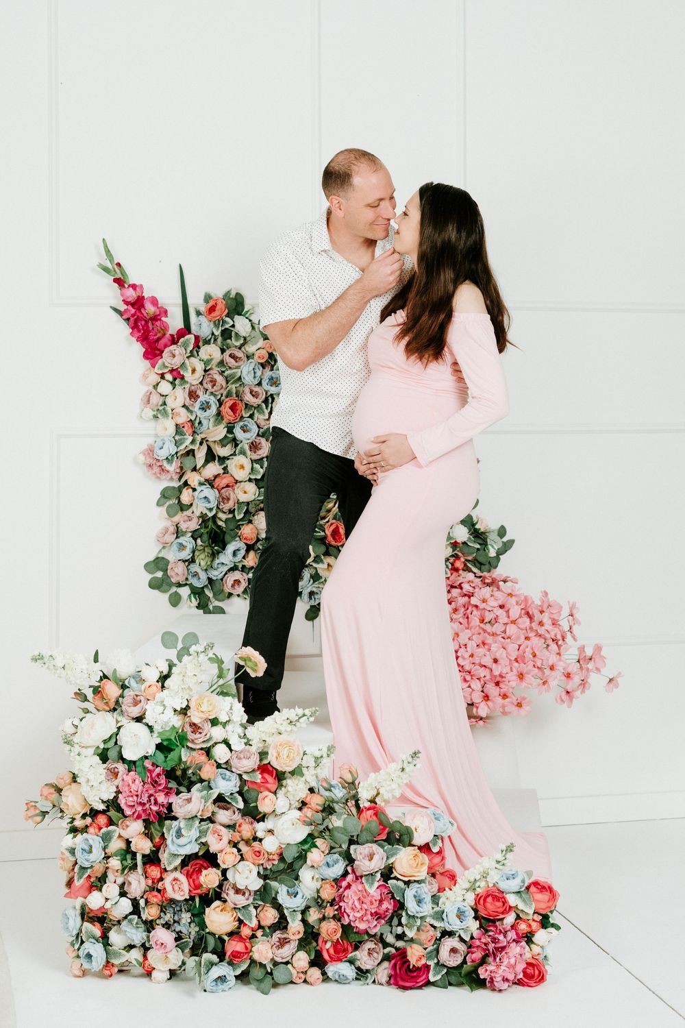 Romantic maternity photoshoot with pink floral arrangements against white backdrop showing couple in intimate poses.
