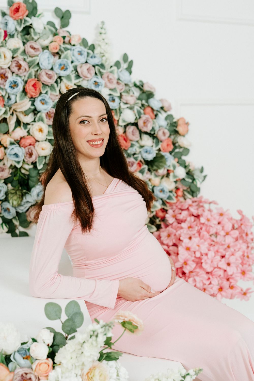 Maternity photo shoot with floral backdrop featuring pink roses and eucalyptus in soft, romantic lighting.