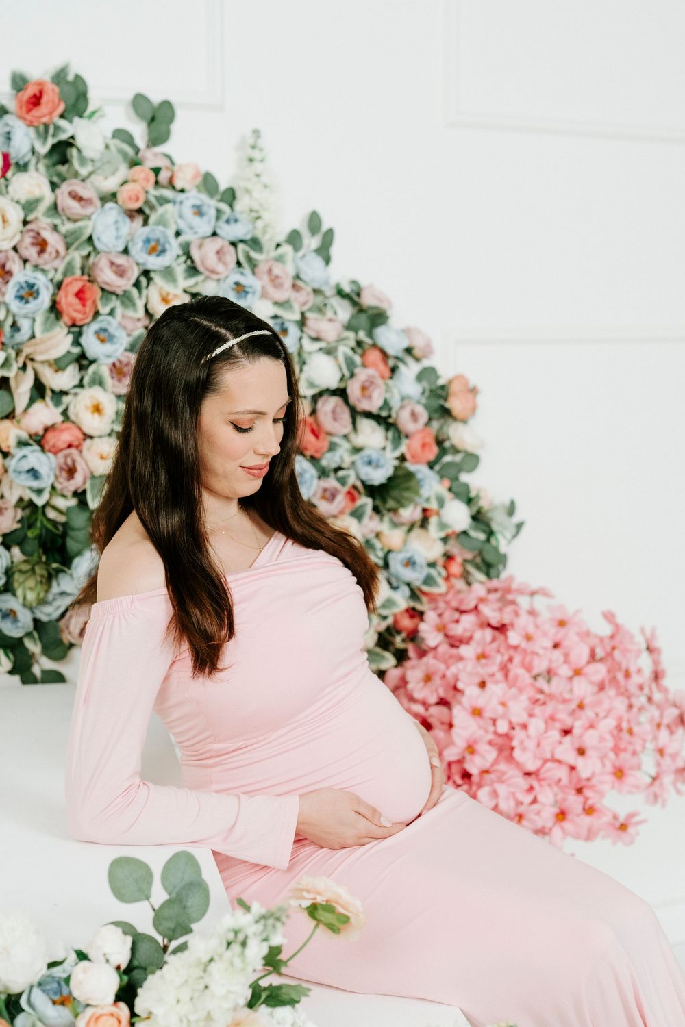 A maternity photo shoot with pink dress against white wall with pink and coral floral decorations and eucalyptus leaves.