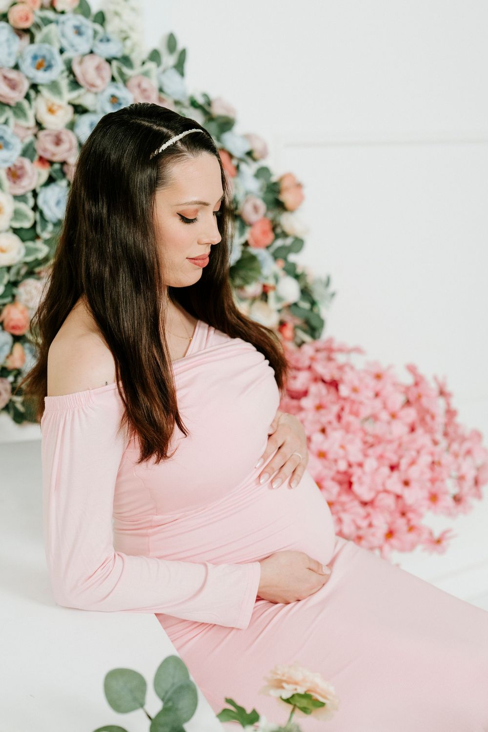 Maternity photo of an expectant mother in a pink off-shoulder dress against a floral backdrop.