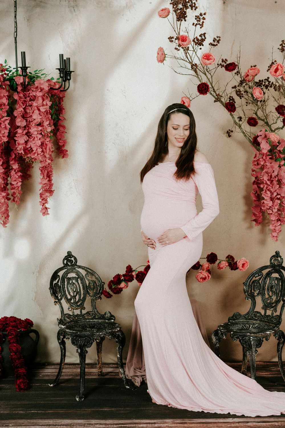 A figure in a flowing pink dress poses gracefully against a wall adorned with pink blossoms and ornate vintage benches.