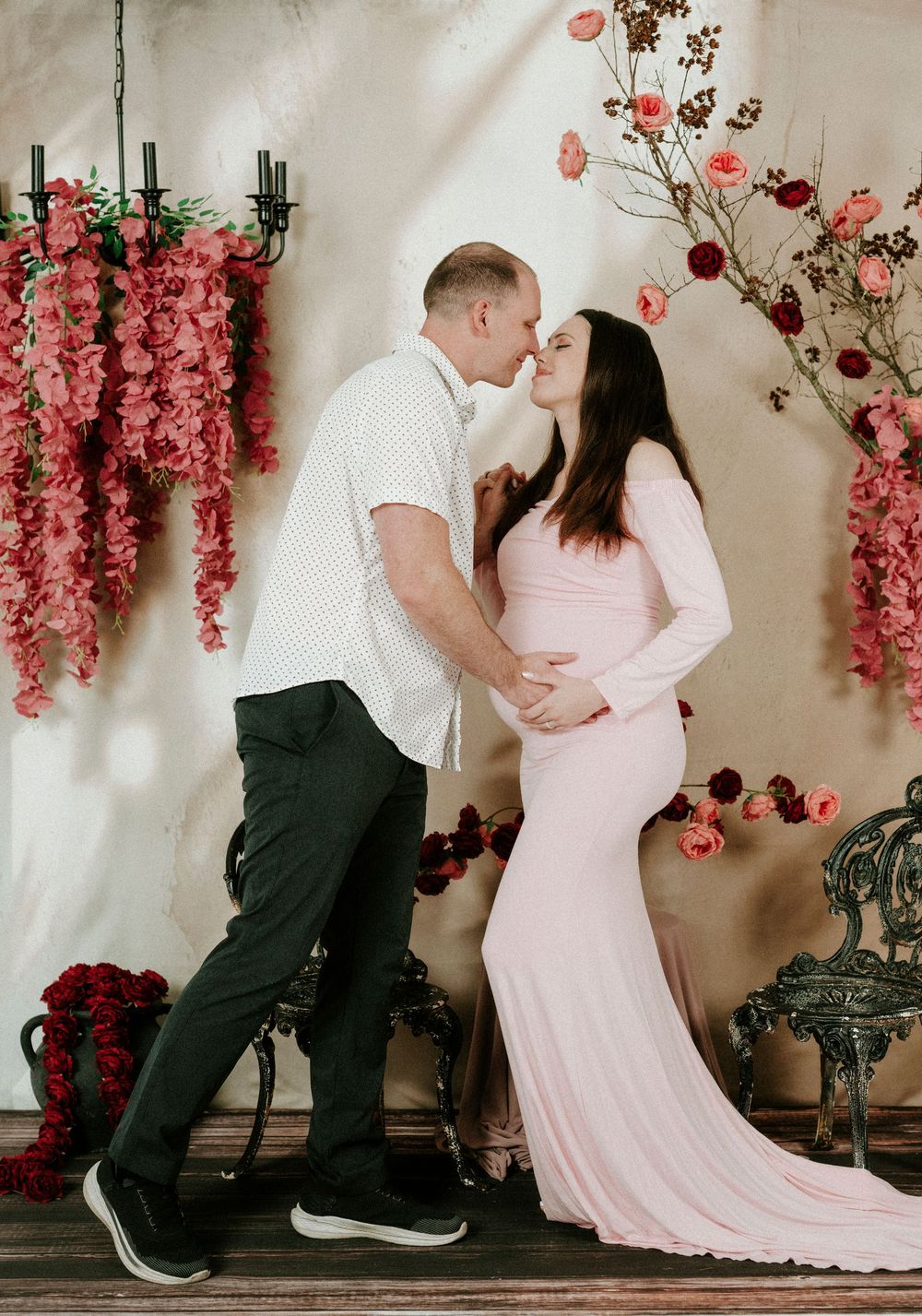 Romantic maternity photo with couple embracing under hanging pink floral arrangements against white backdrop.