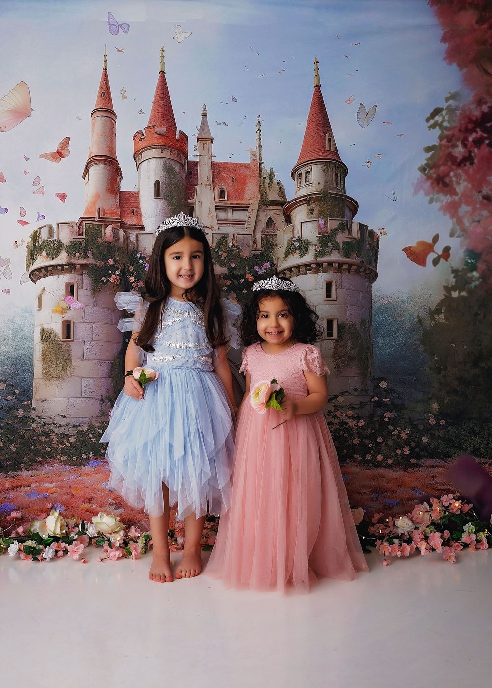 Two children in pastel dresses pose together against a fairytale castle backdrop with flowers.