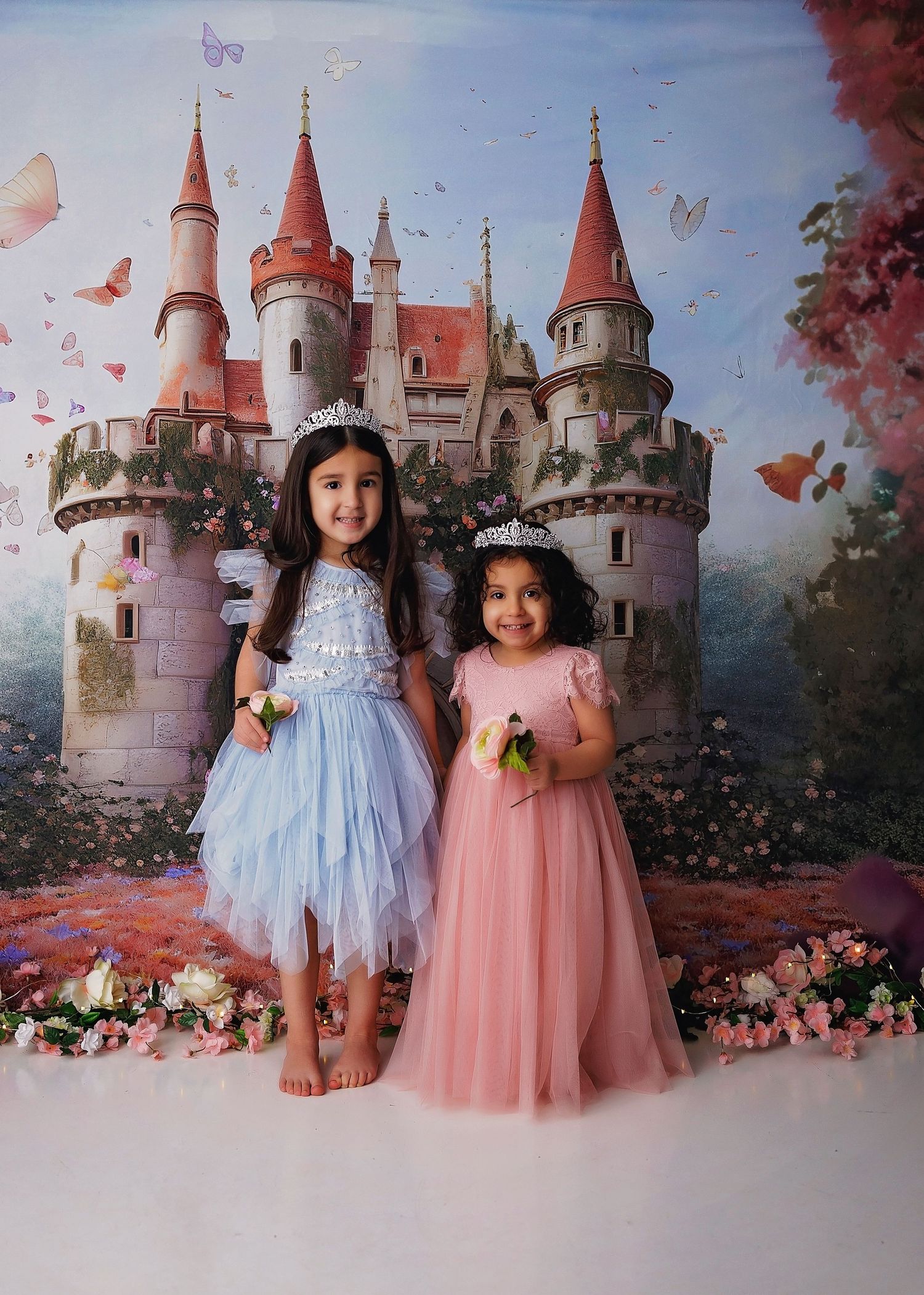 Two children in pastel dresses pose together against a fairytale castle backdrop with flowers.