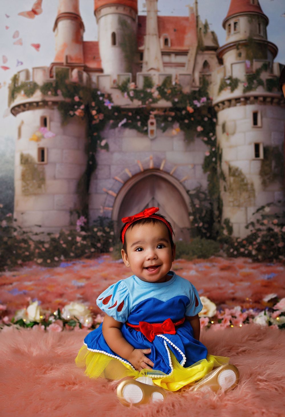 Sweet baby in Snow White costume smiles while sitting against castle backdrop in professional photo studio setting.