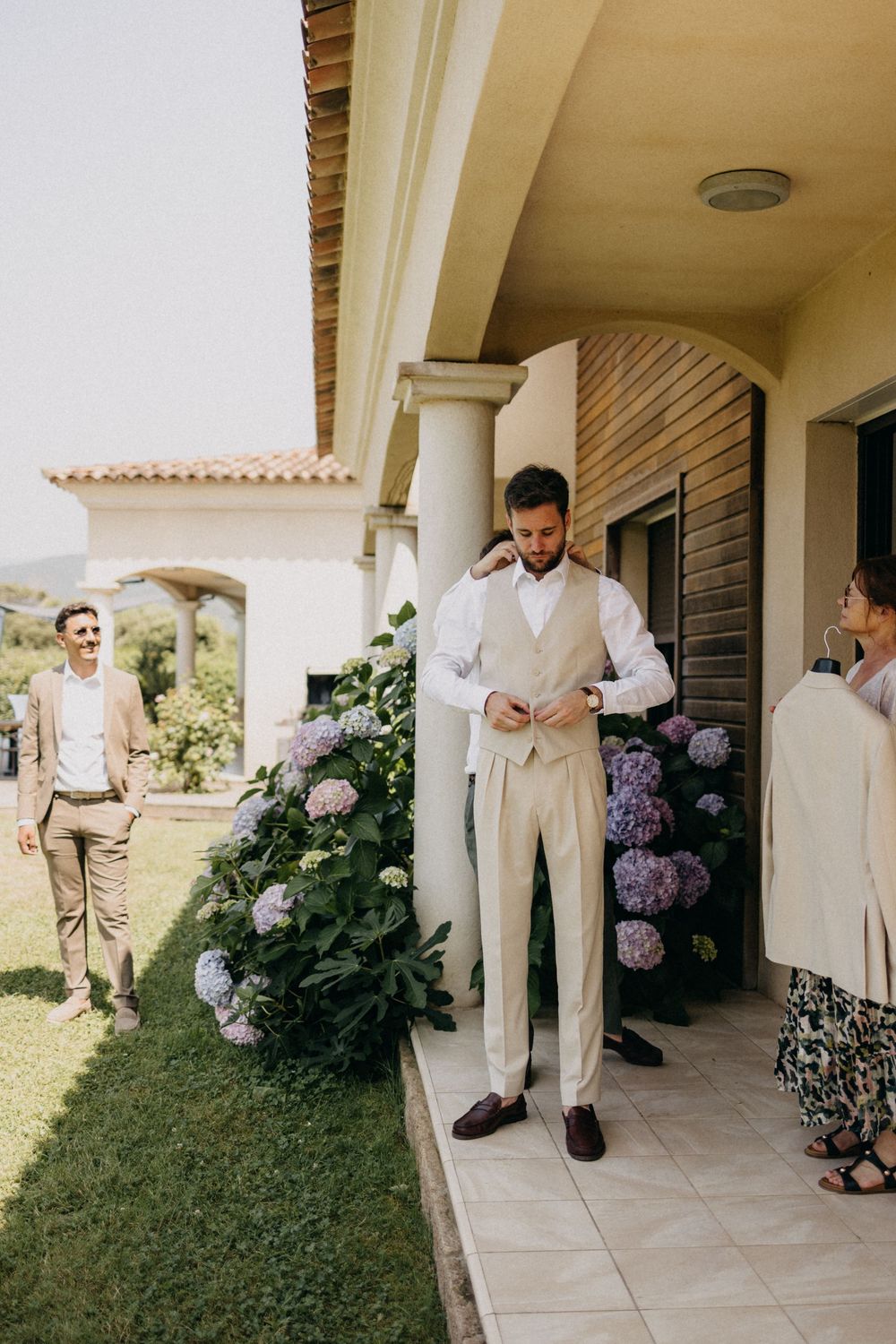 Wedding party members in light colored suits standing on a covered porch with purple flowers.