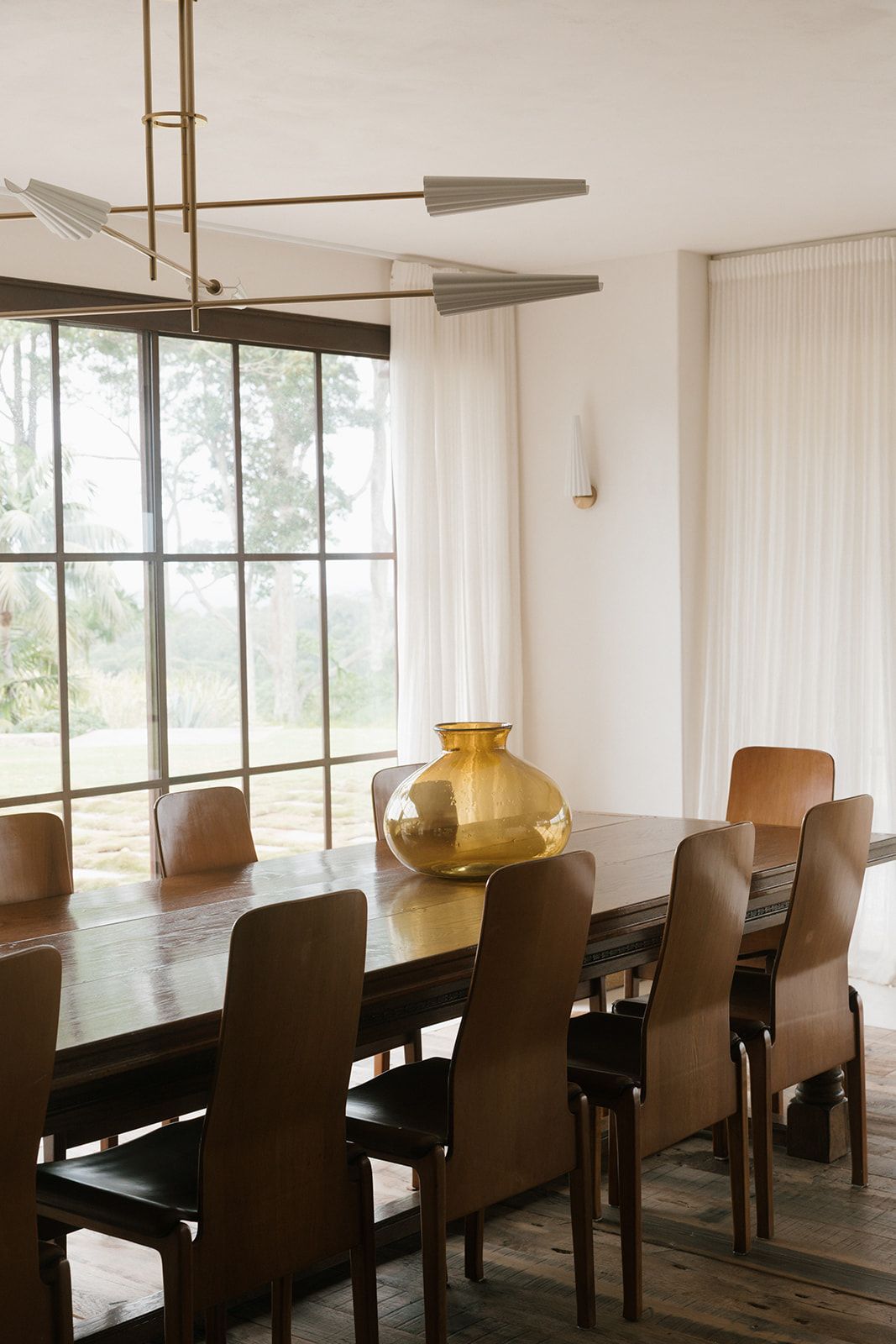 Modern dining room with dark wood table, brown chairs, and large windows overlooking trees in natural light.