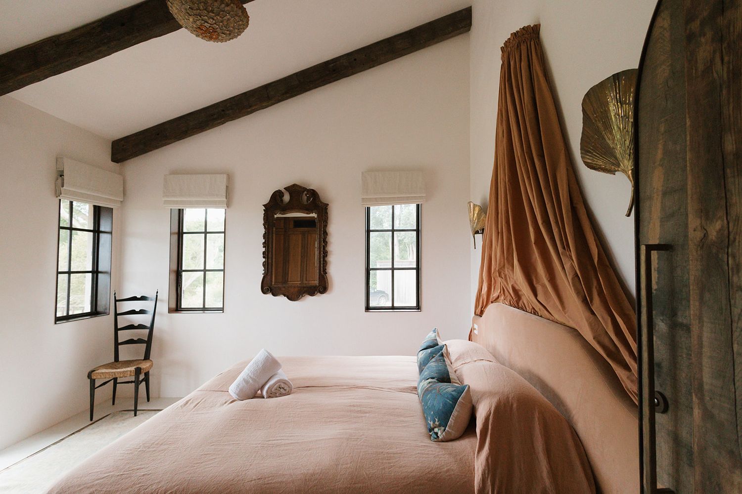 A serene rustic bedroom interior with wooden beams, white walls, and multiple windows letting in natural light.