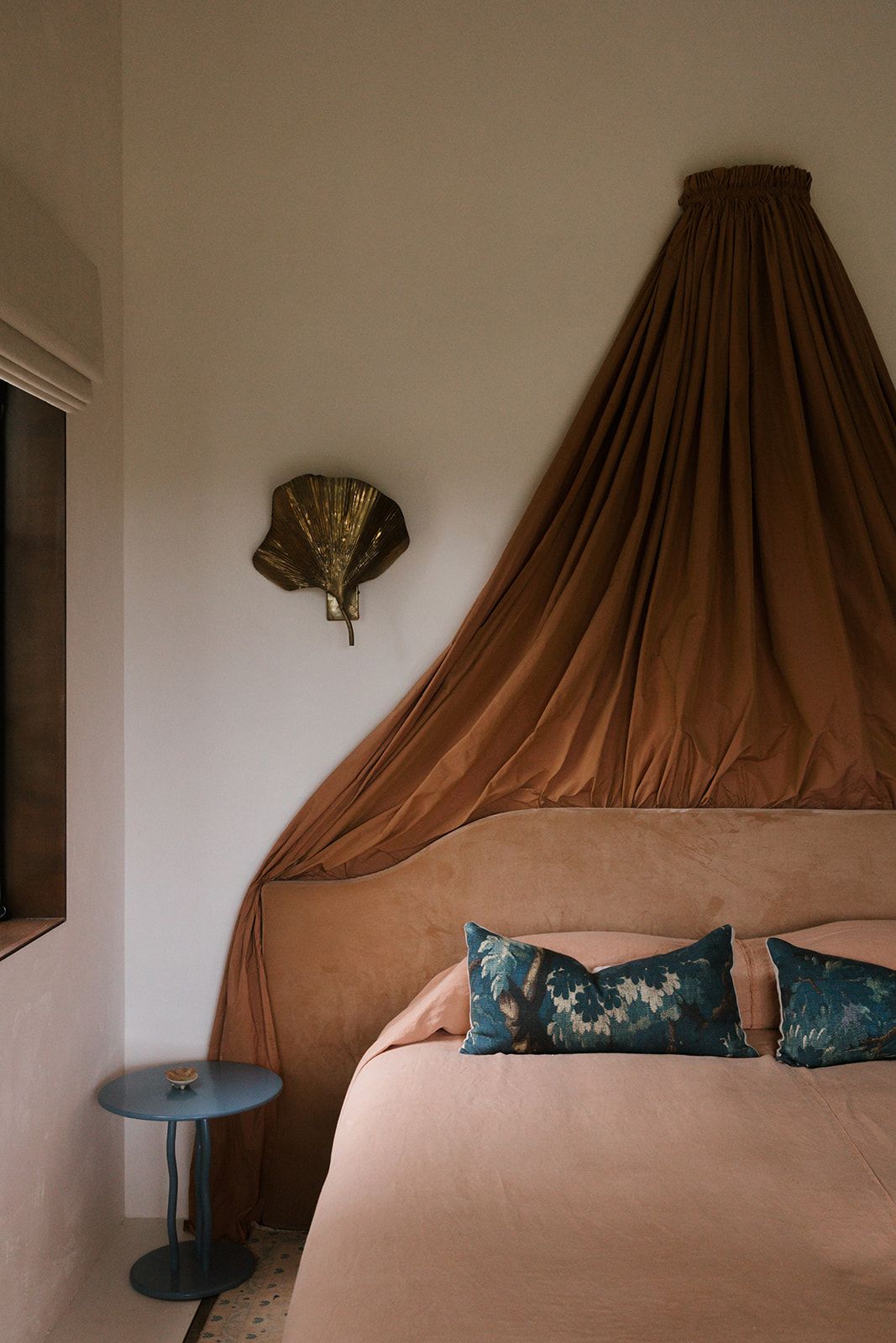 Modern bedroom with brown draped canopy, beige bedding, teal accent pillows, and decorative gold fan on wall.
