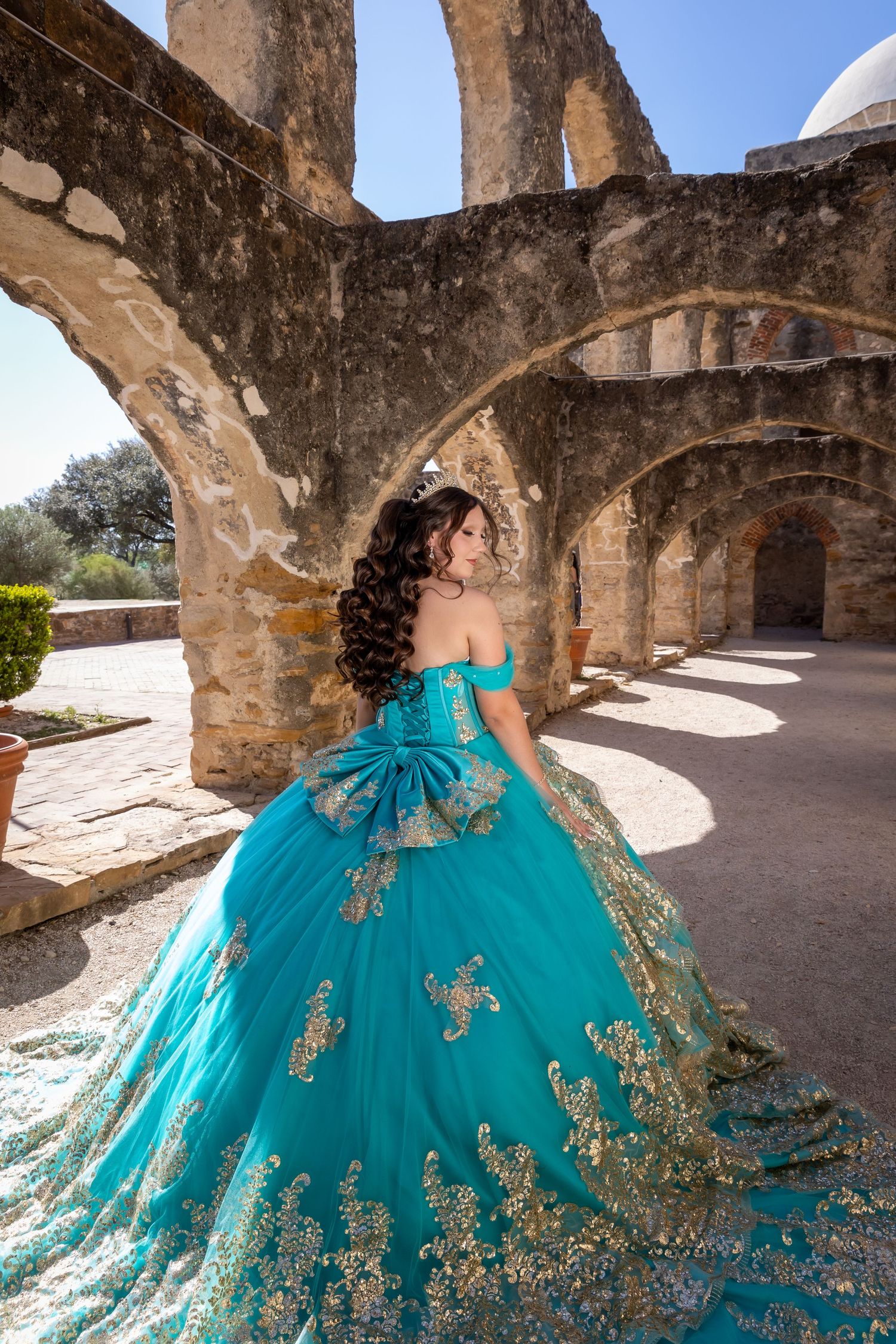 A burgundy quinceañera dress with gold embroidery details is showcased in an outdoor garden setting on stone steps.