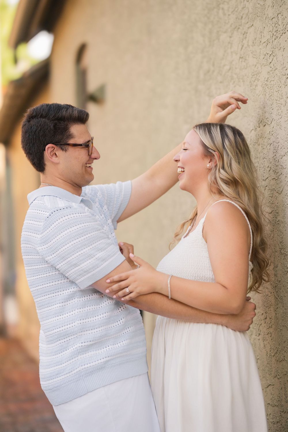 A couple in white outfits shares a joyful candid moment together against a neutral wall outdoors.