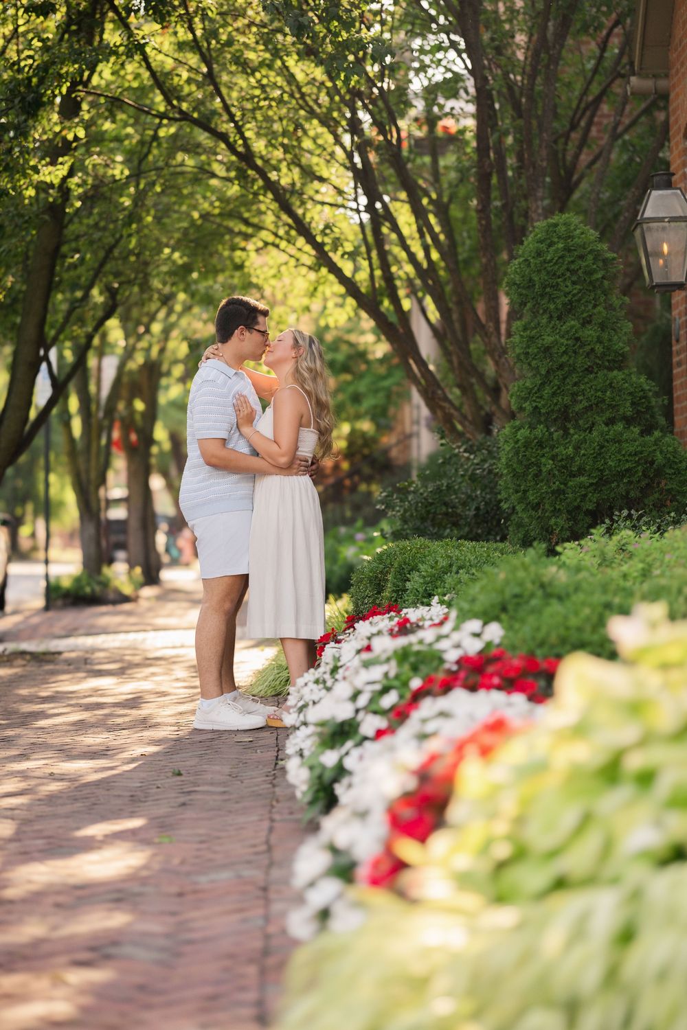 A romantic couple shares a tender moment on a brick path colorful flowers and lush trees German Village Columbus Ohio Engagement Session.