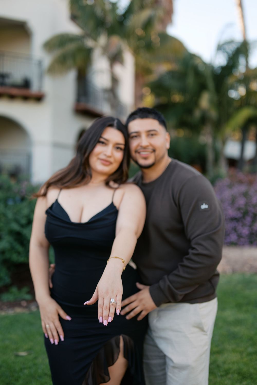 A couple poses together outdoors near palm trees and purple flowers at a resort-style property.