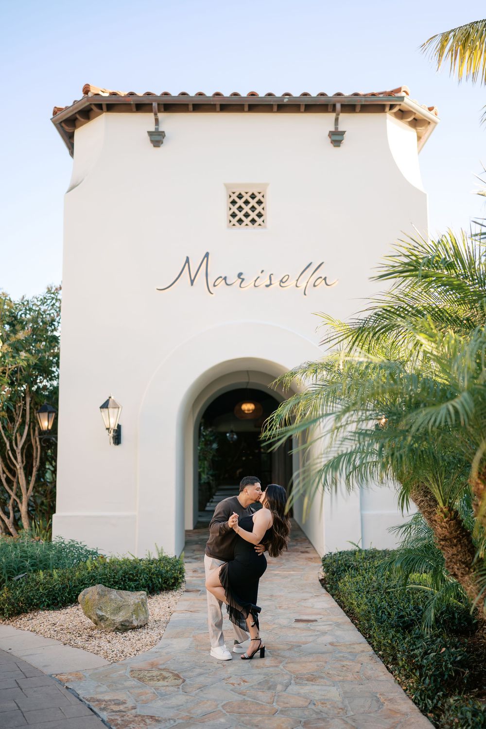 A couple embraces in front of a Mediterranean-style building entrance with 'Marisella' signage and tropical landscaping.