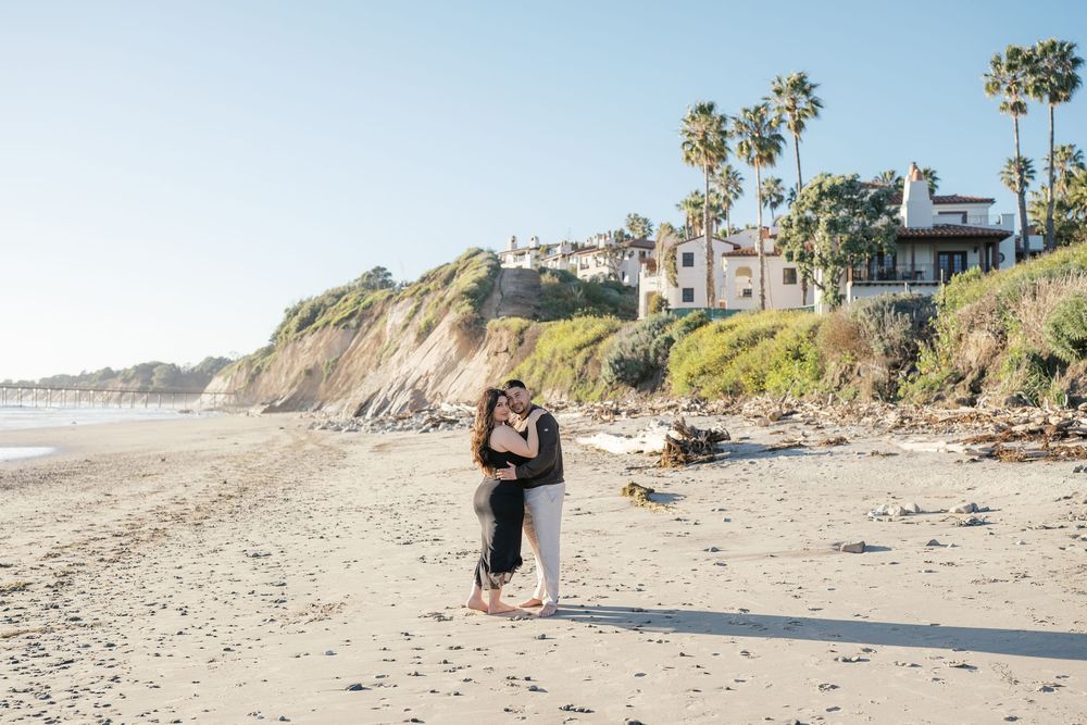 A couple shares a romantic moment on a scenic beach with coastal cliffs and palm trees in the background.