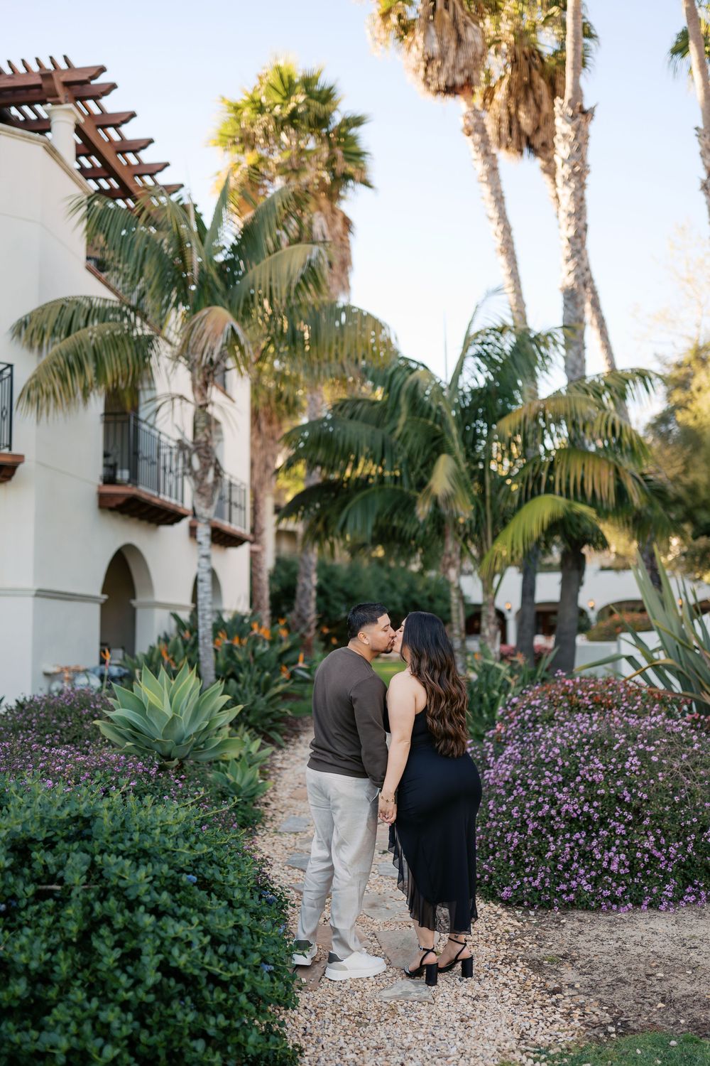 A romantic couple strolls through a tropical garden path lined with palm trees and purple flowers.