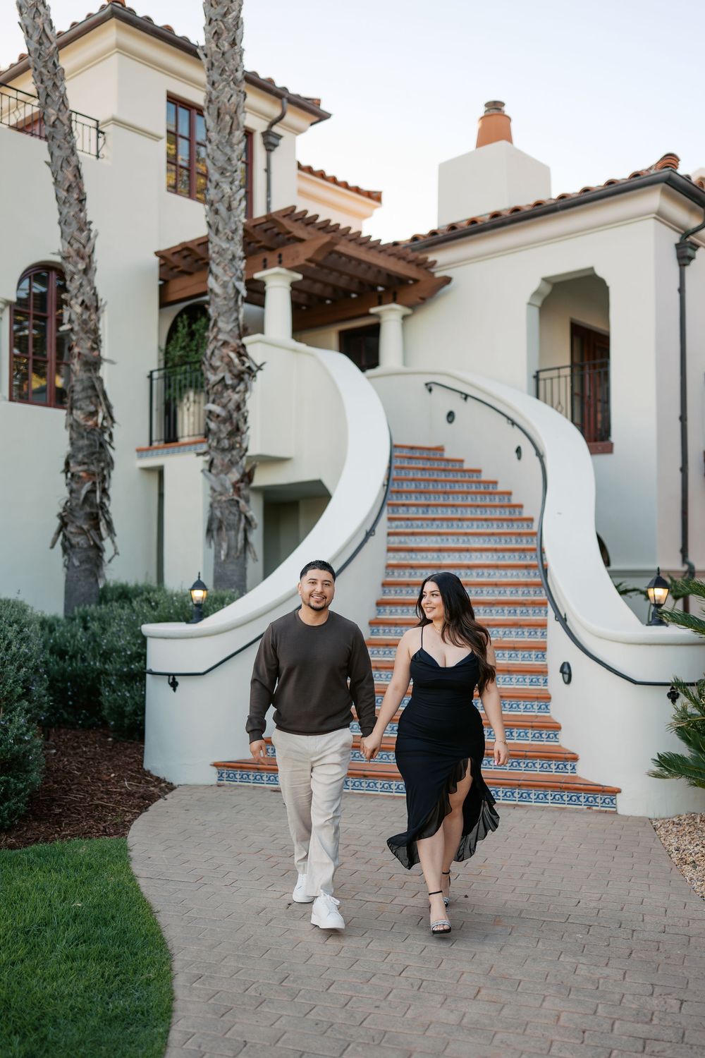 Couple walking together on a curved pathway leading to Spanish-style stairs with white stucco walls and palm trees.