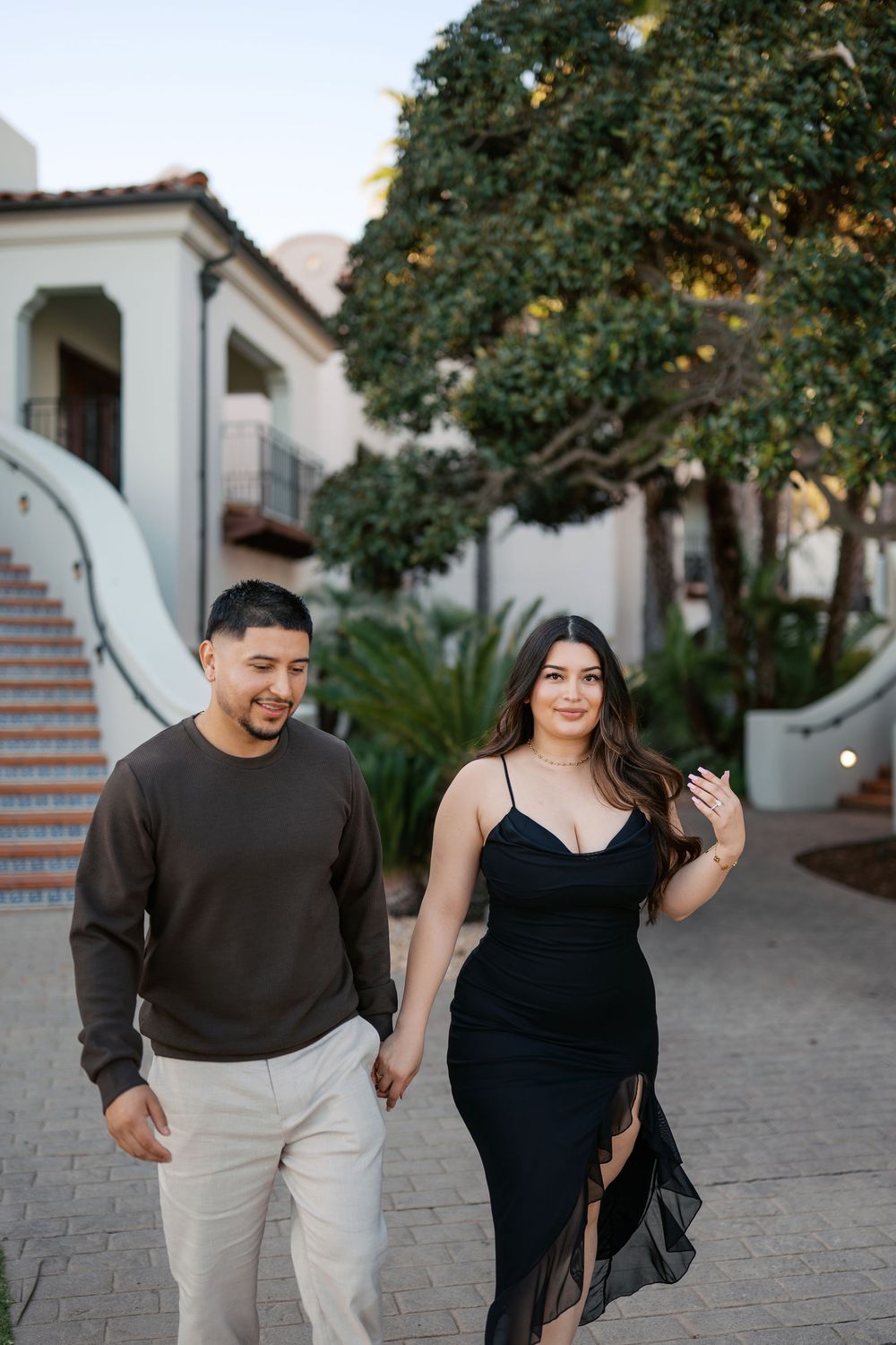 A couple walks and laughs together on a paved path outside a Spanish-style building with arched doorways.