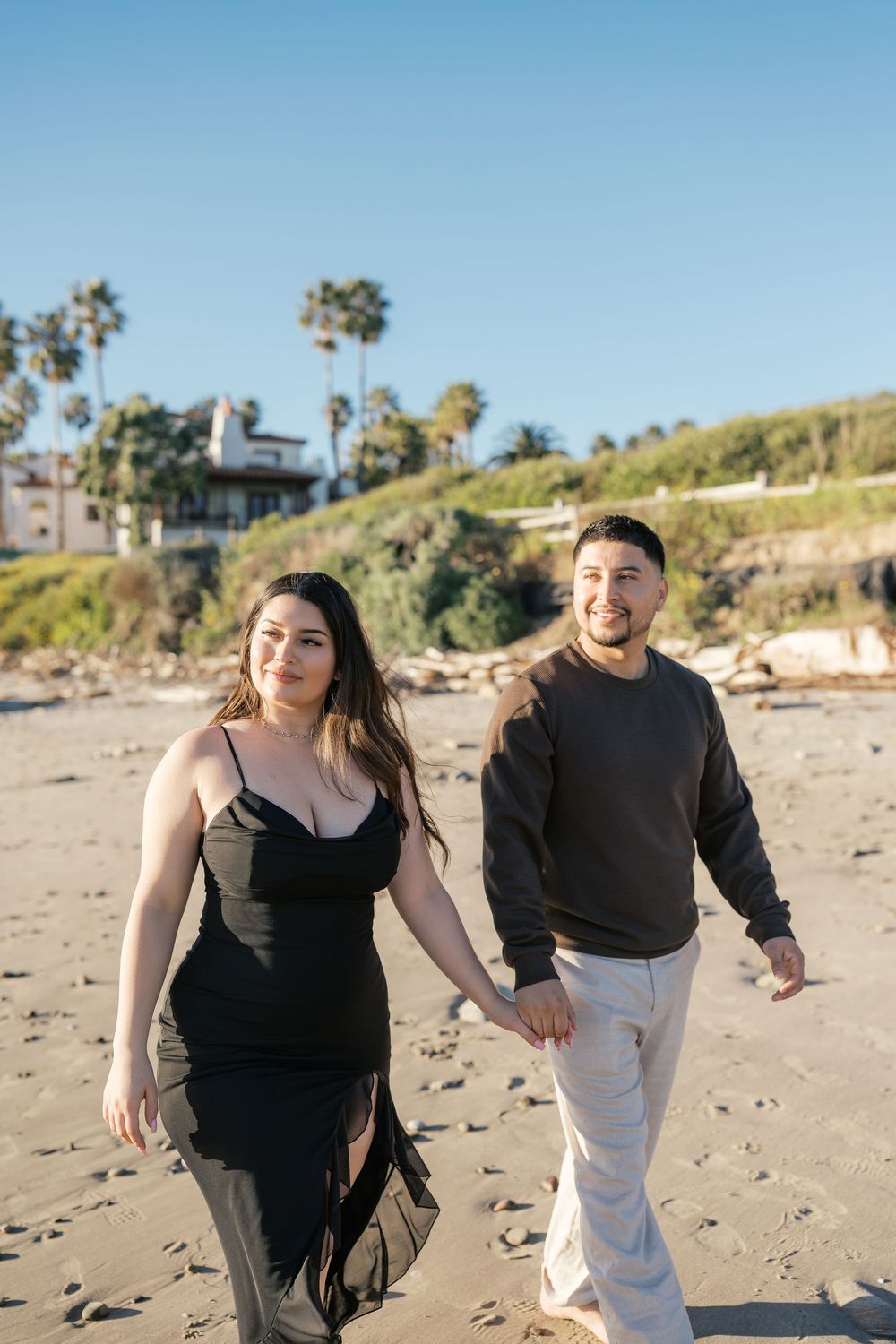 A couple in black and white outfits playfully interacts while walking on a sunny beach with palm trees in the background.